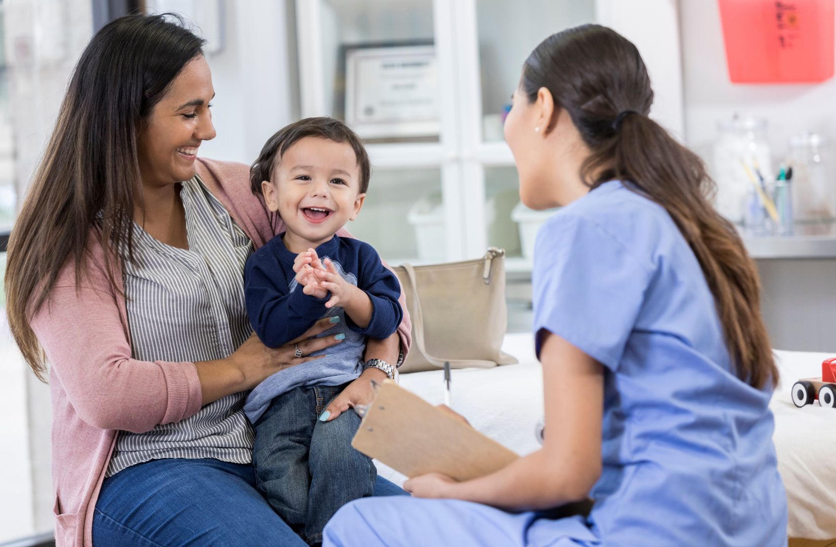 A smiling mother and toddler son sits in an exam room and listen to a healthcare professional.